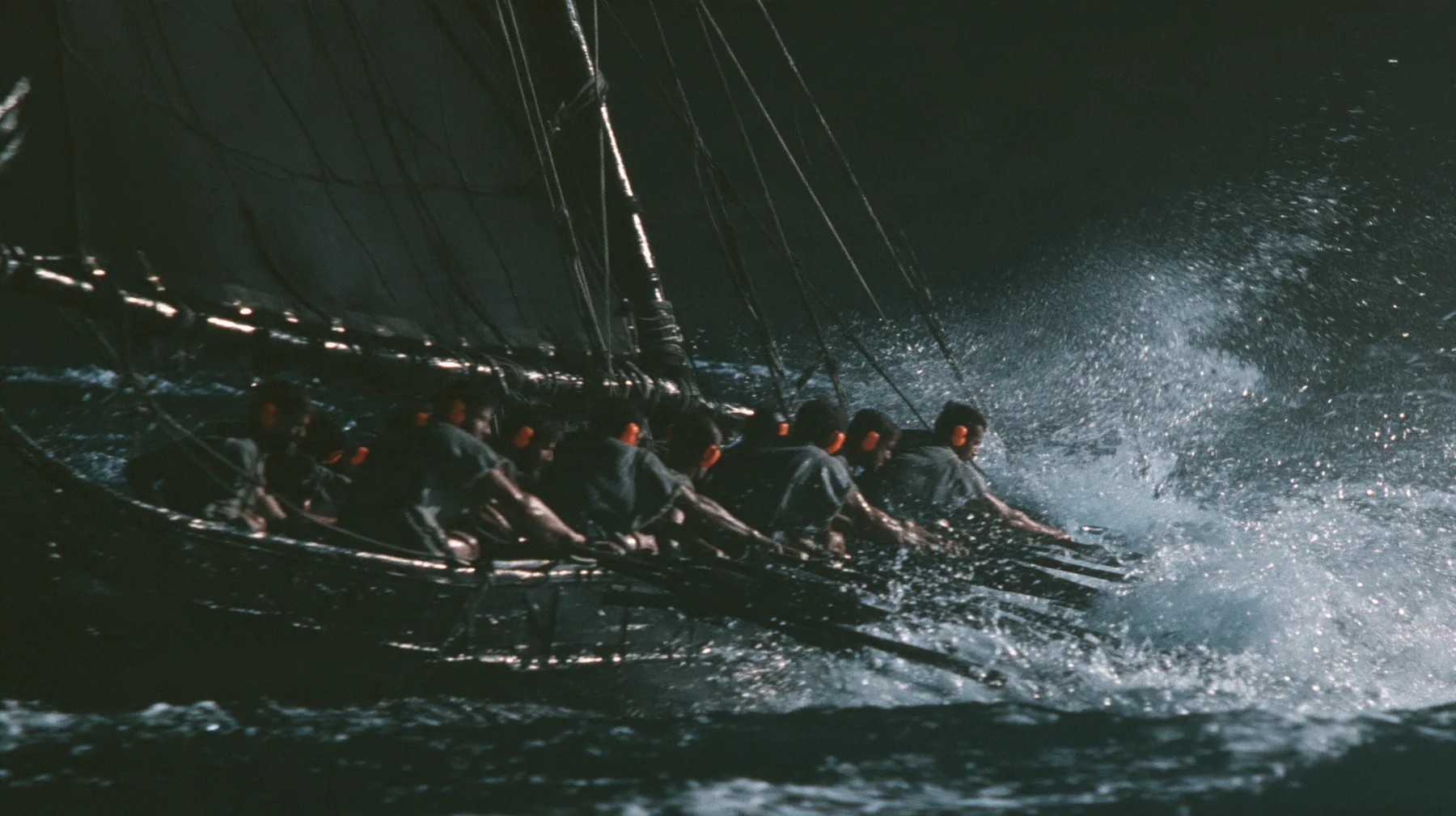 medium shot over the gunwale of a long black ancient Greek galley with a single mast, a square sail and two banks of oars, rowers in undyed linen tunics pulling the oars in time, each with bright orange foam earplugs…