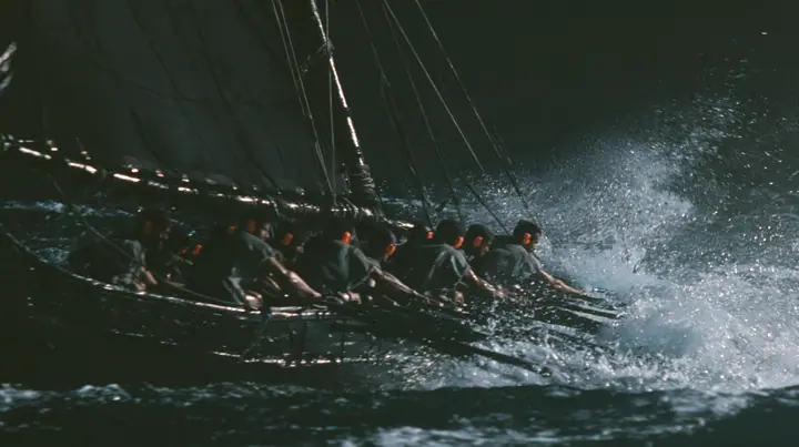 medium shot over the gunwale of a long black ancient Greek galley with a single mast, a square sail and two banks of oars, rowers in undyed linen tunics pulling the oars in time, each with bright orange foam earplugs…