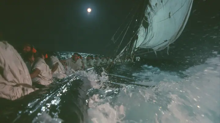 medium shot over the gunwale of a long black ancient Greek galley with a single mast, a square sail and two banks of oars, rowers in undyed linen tunics pulling the oars in time, each with bright orange foam earplugs…