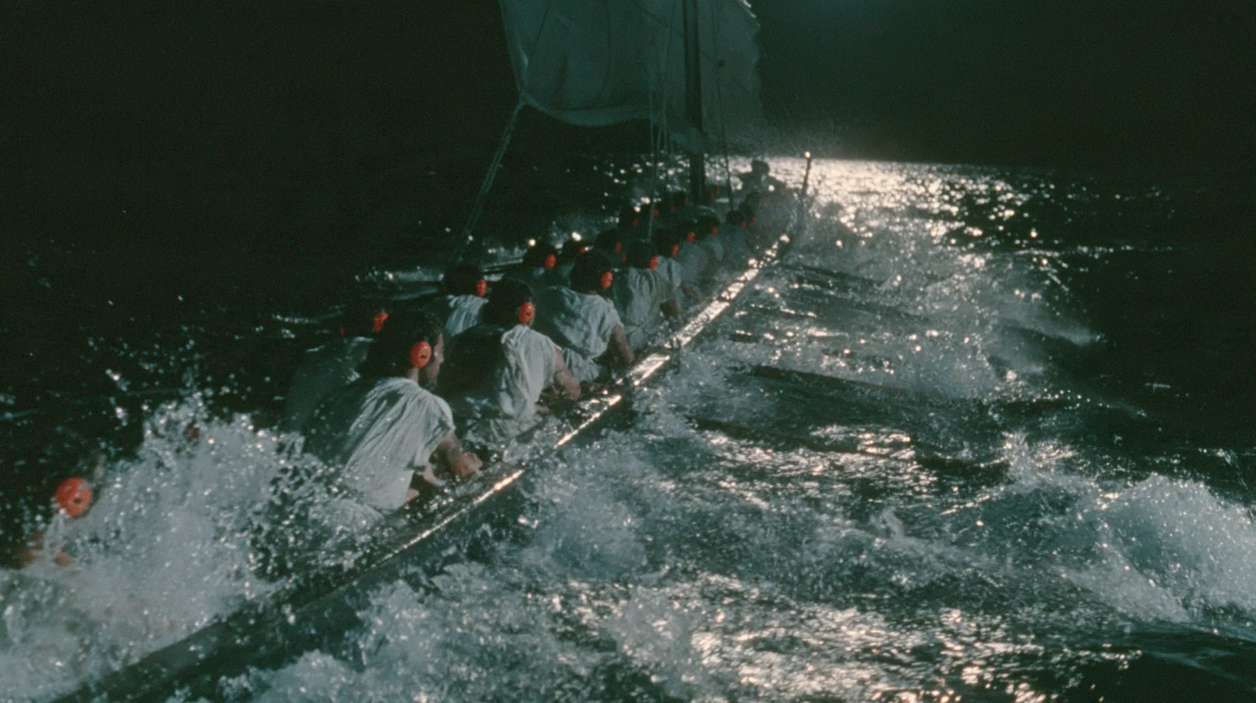 medium shot over the gunwale of a long black ancient Greek galley with a single mast, a square sail and two banks of oars, rowers in undyed linen tunics pulling the oars in time, each with bright orange foam earplugs…