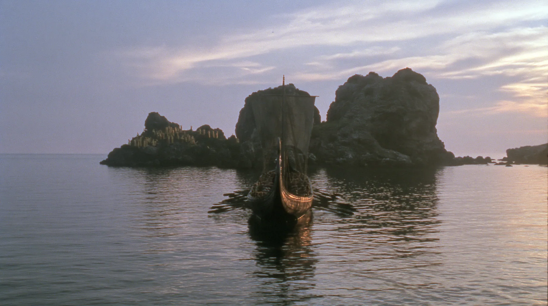 extreme wide shot from behind a long black ancient Greek galley with a single mast, a square sail and two banks of oars on a glassy sea, on the horizon a cluster of black rocks with tiny gold-lit figures of women,…