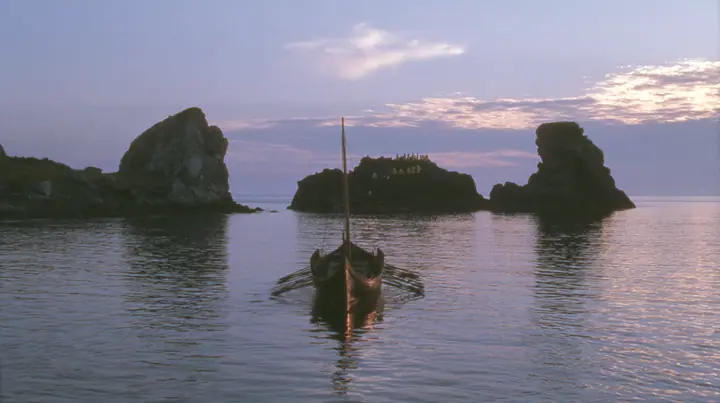 extreme wide shot from behind a long black ancient Greek galley with a single mast, a square sail and two banks of oars on a glassy sea, on the horizon a cluster of black rocks with tiny gold-lit figures of women,…