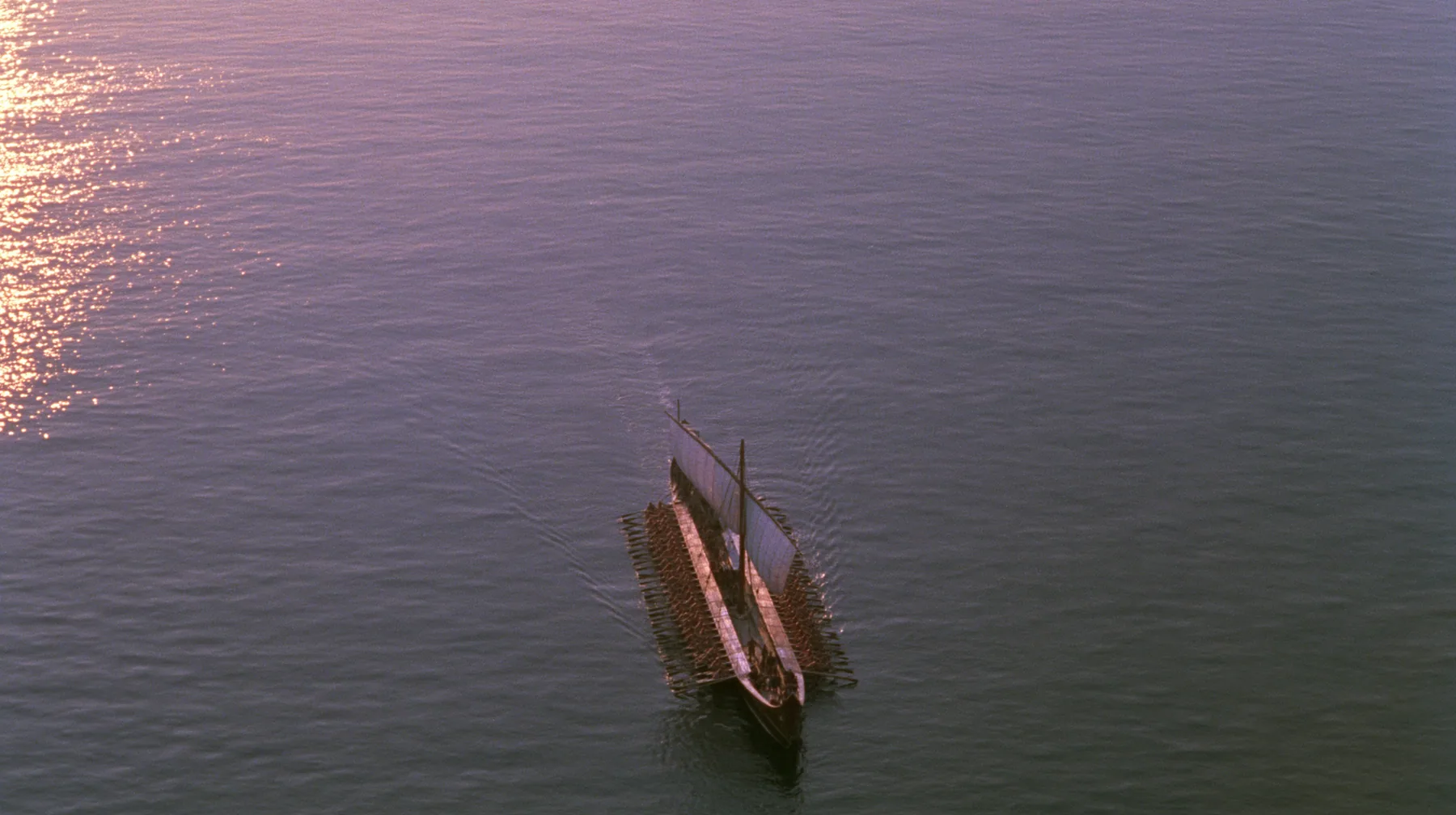 straight-down aerial of a long black ancient Greek galley with a single mast, a square sail and two banks of oars at dusk on a glassy violet sea, the two rows of oars in perfect order like lines on a page, dusk, low…