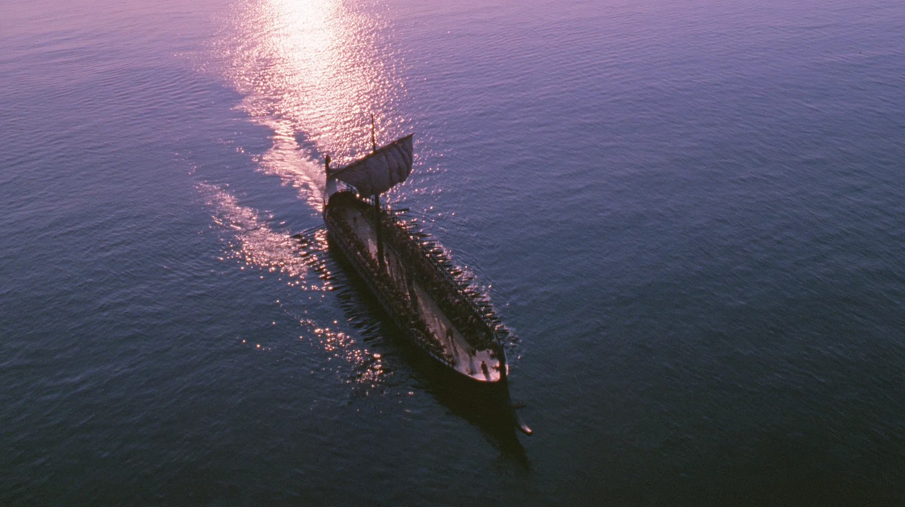 straight-down aerial of a long black ancient Greek galley with a single mast, a square sail and two banks of oars at dusk on a glassy violet sea, the two rows of oars in perfect order like lines on a page, dusk, low…