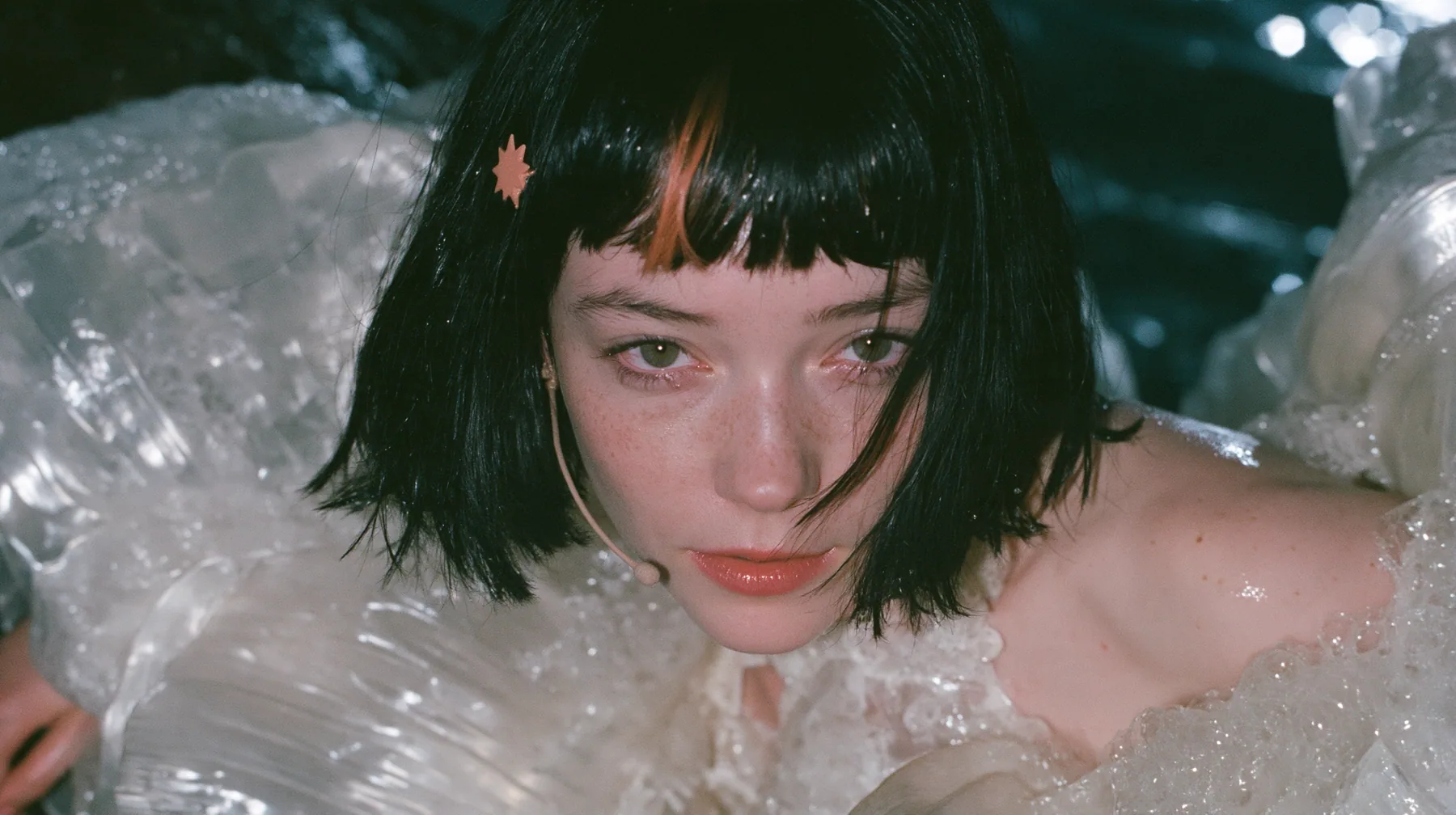close-up of a Caucasian American woman in her mid-twenties with pale skin and light freckles, a glossy black blunt jaw-length bob with heavy straight bangs and one clay-orange streak through the bangs, a small flat…