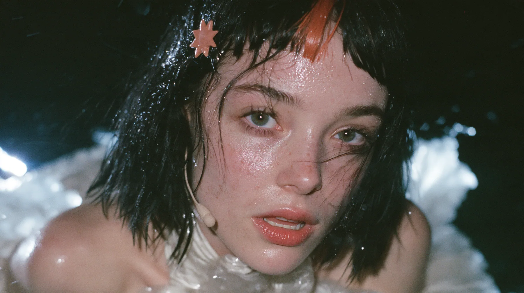 close-up of a Caucasian American woman in her mid-twenties with pale skin and light freckles, a glossy black blunt jaw-length bob with heavy straight bangs and one clay-orange streak through the bangs, a small flat…