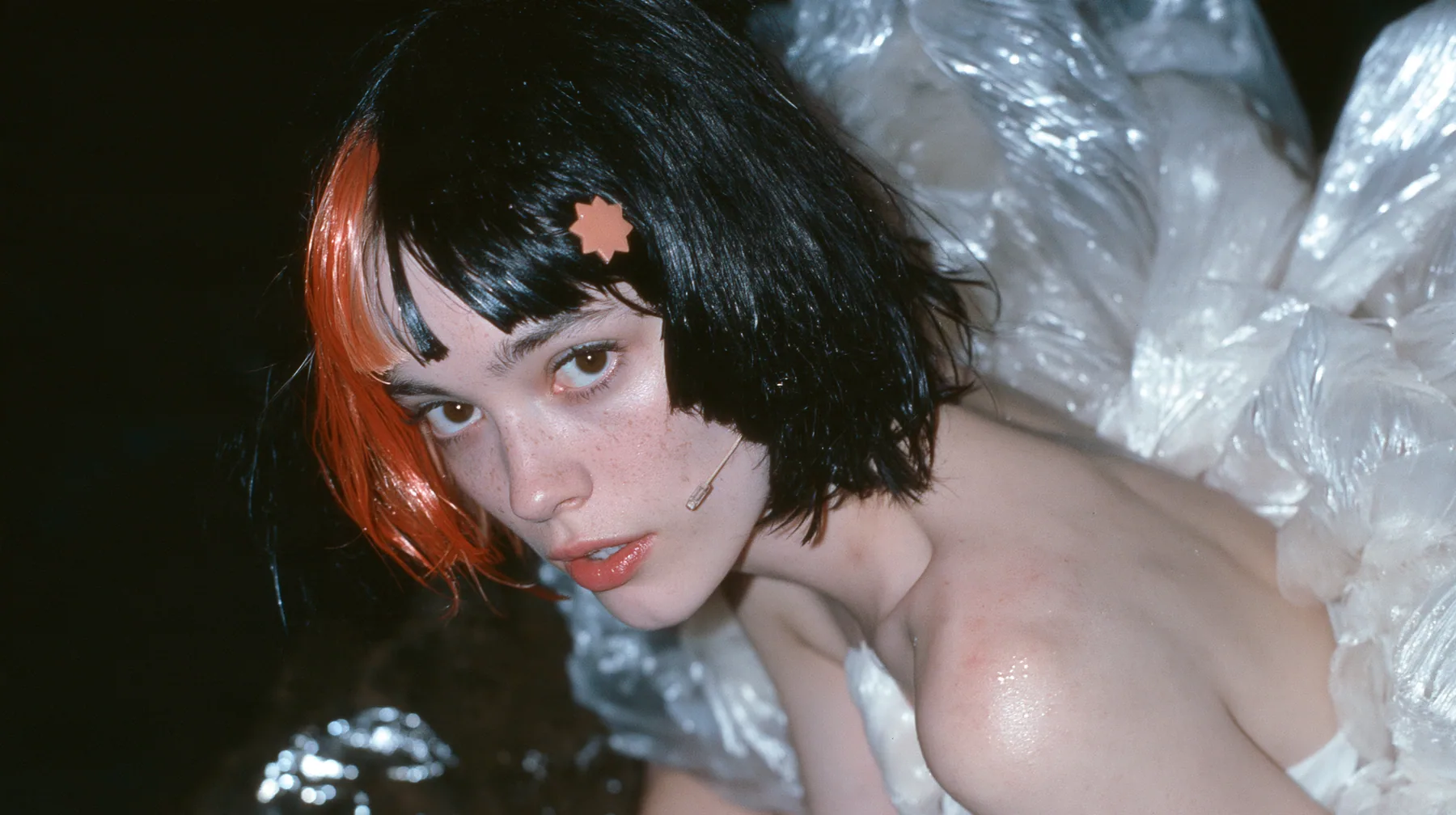 medium close-up of a Caucasian American woman in her mid-twenties with pale skin and light freckles, a glossy black blunt jaw-length bob with heavy straight bangs and one clay-orange streak through the bangs, a small…