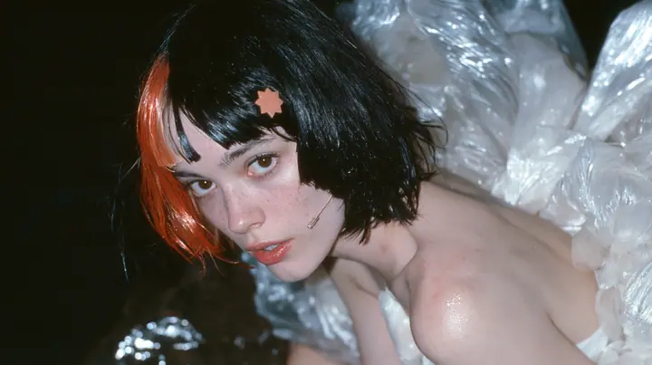 medium close-up of a Caucasian American woman in her mid-twenties with pale skin and light freckles, a glossy black blunt jaw-length bob with heavy straight bangs and one clay-orange streak through the bangs, a small…