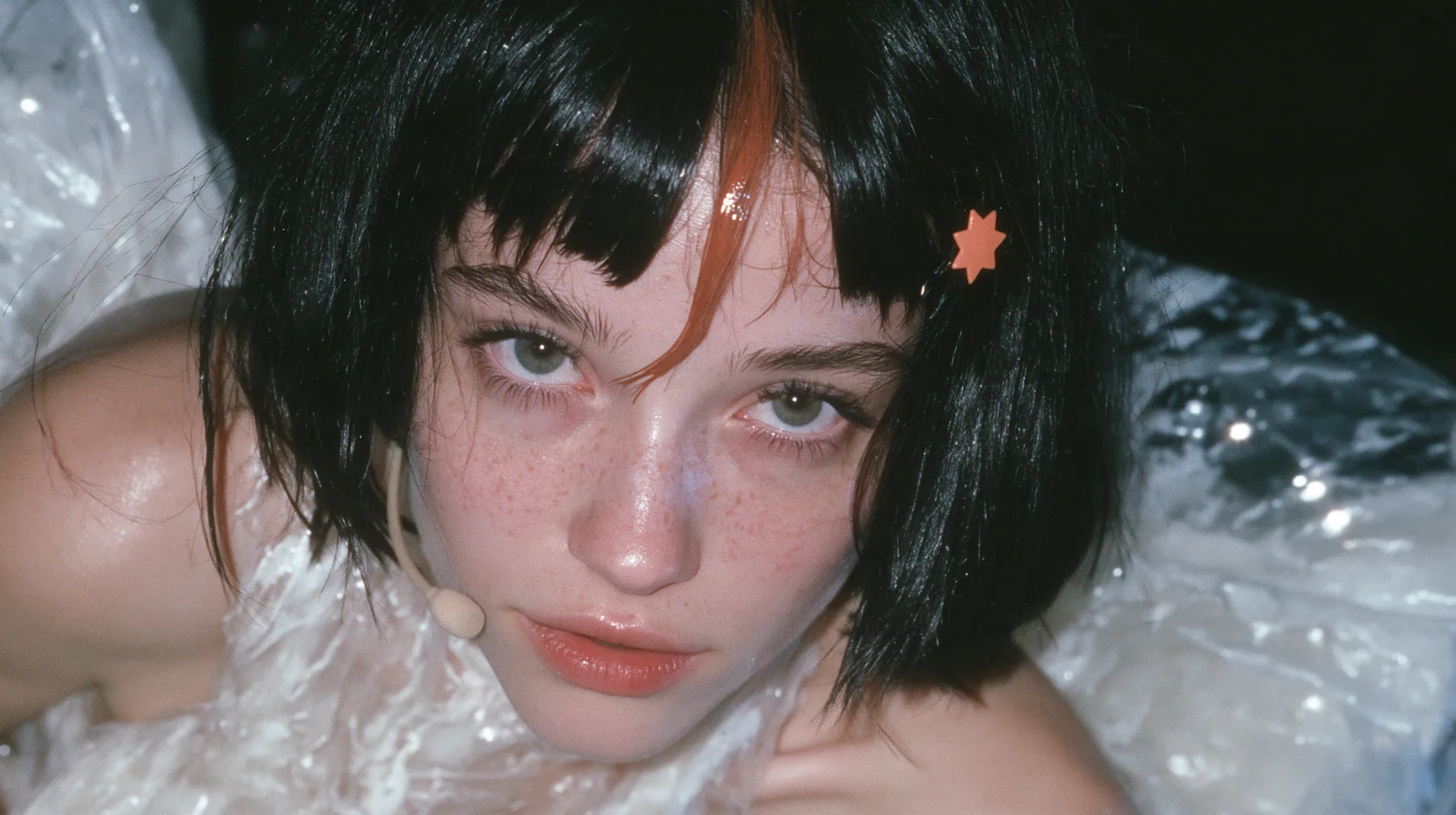 close-up of a Caucasian American woman in her mid-twenties with pale skin and light freckles, a glossy black blunt jaw-length bob with heavy straight bangs and one clay-orange streak through the bangs, a small flat…