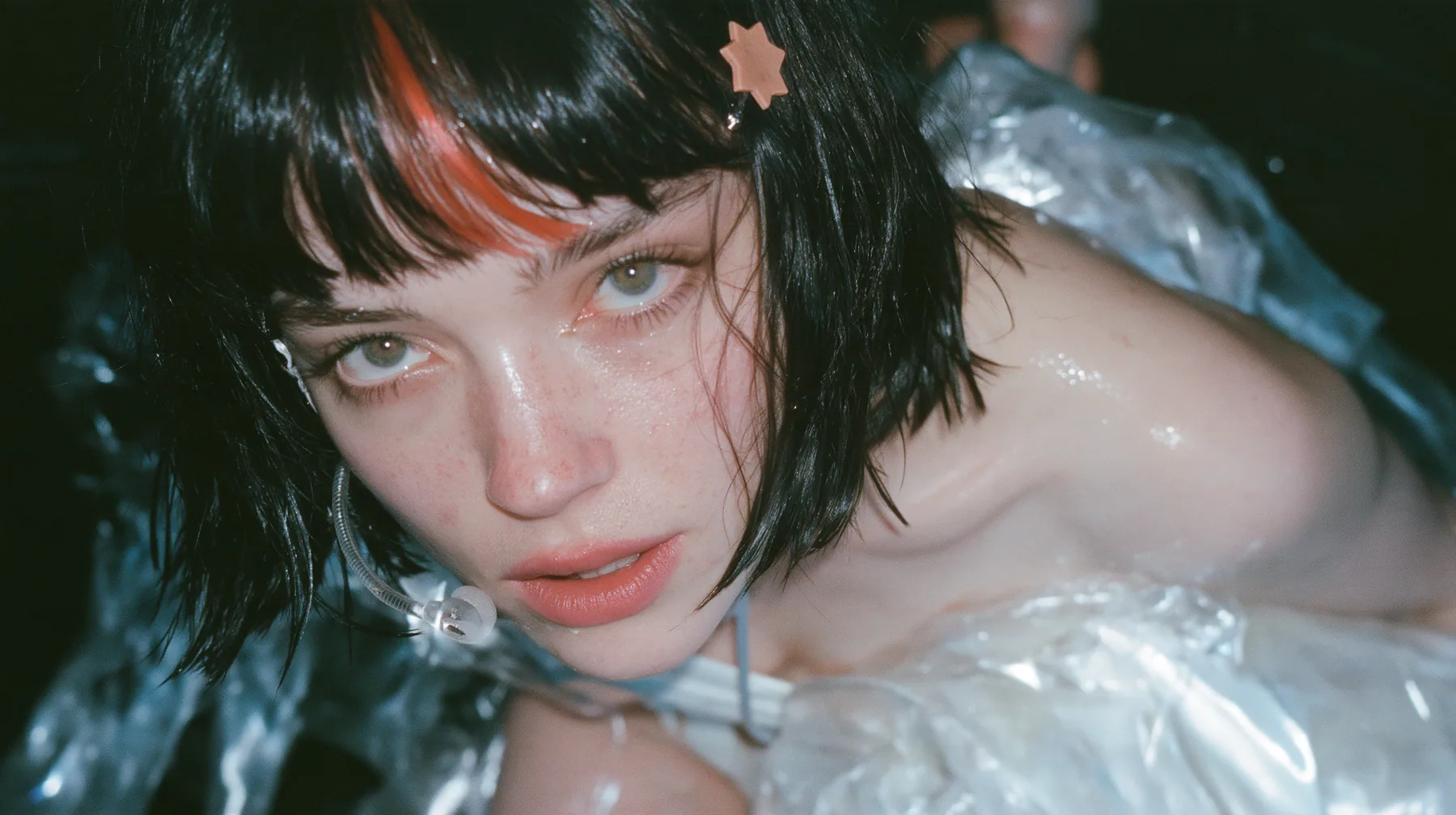 close-up of a Caucasian American woman in her mid-twenties with pale skin and light freckles, a glossy black blunt jaw-length bob with heavy straight bangs and one clay-orange streak through the bangs, a small flat…