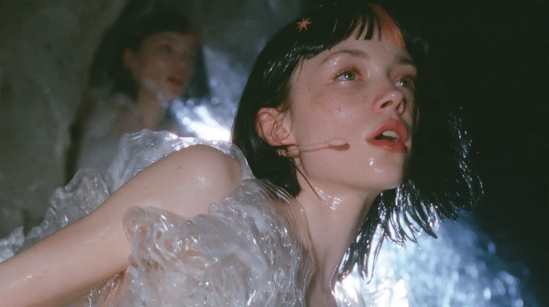 medium shot of a Caucasian American woman in her mid-twenties with pale skin and light freckles, a glossy black blunt jaw-length bob with heavy straight bangs and one clay-orange streak through the bangs, a small…