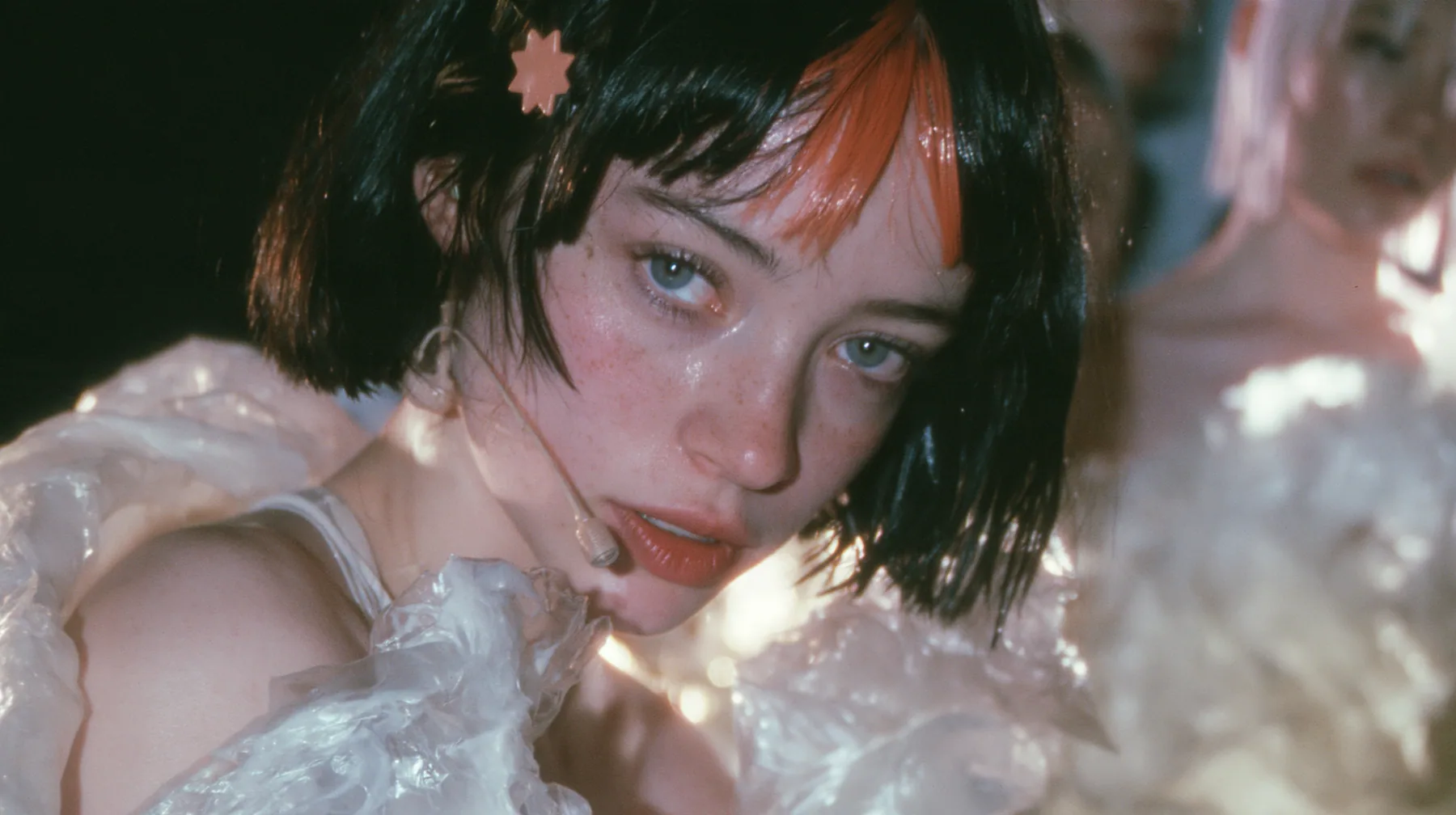 medium shot of a Caucasian American woman in her mid-twenties with pale skin and light freckles, a glossy black blunt jaw-length bob with heavy straight bangs and one clay-orange streak through the bangs, a small…