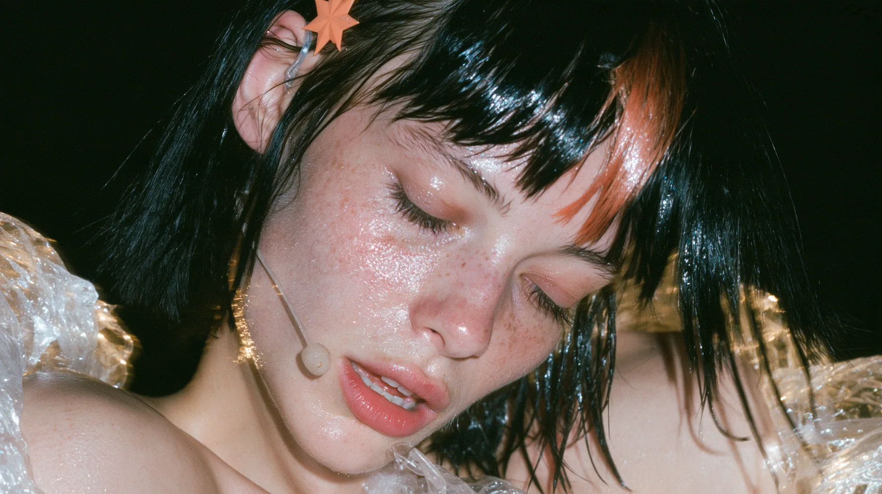 close-up of a Caucasian American woman in her mid-twenties with pale skin and light freckles, a glossy black blunt jaw-length bob with heavy straight bangs and one clay-orange streak through the bangs, a small flat…