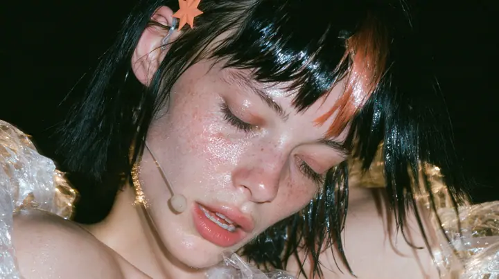 close-up of a Caucasian American woman in her mid-twenties with pale skin and light freckles, a glossy black blunt jaw-length bob with heavy straight bangs and one clay-orange streak through the bangs, a small flat…