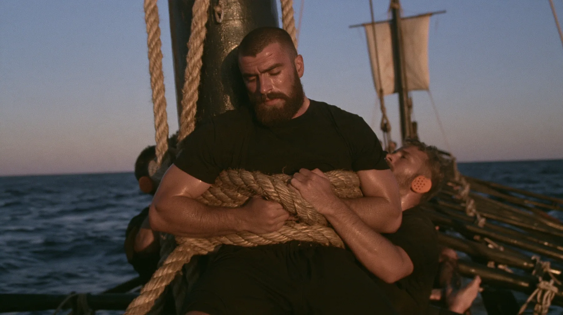 medium shot of a broad-shouldered Caucasian man in his early thirties with a square jaw, a short dark beard, a dark buzz cut and heavy brows, the build of a man who lifts, in a plain black t-shirt and black trousers,…