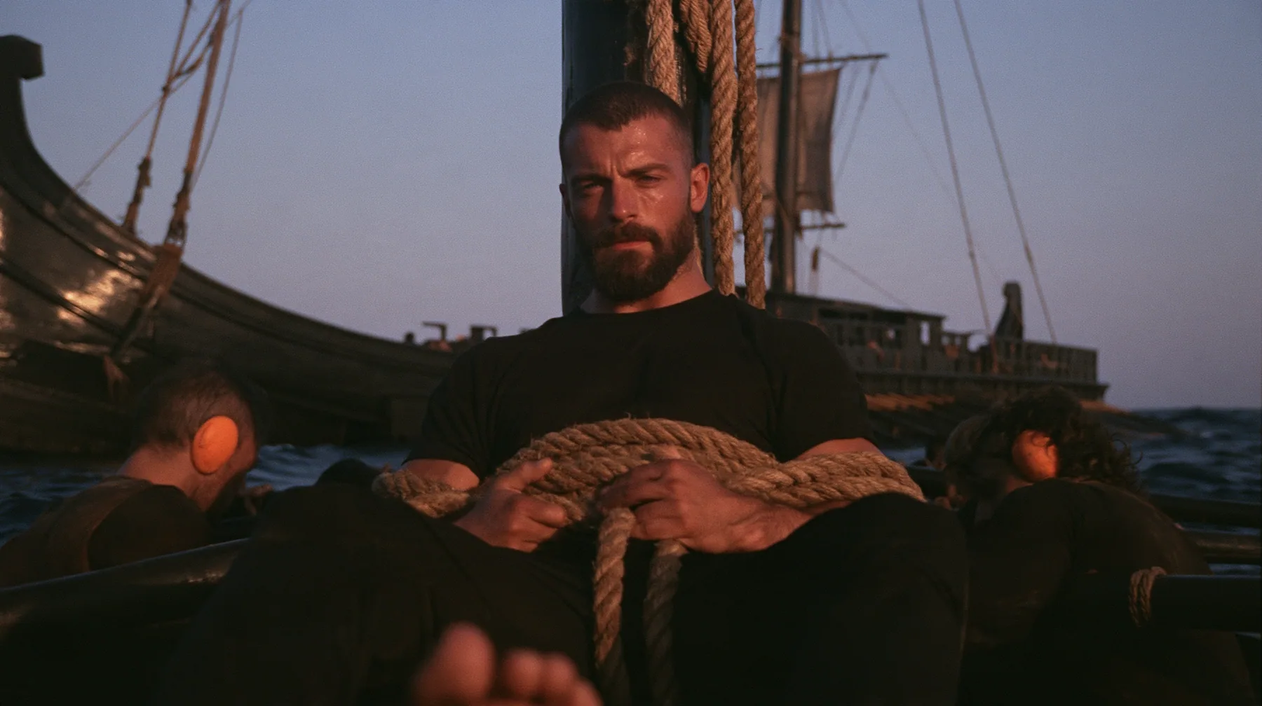 medium shot of a broad-shouldered Caucasian man in his early thirties with a square jaw, a short dark beard, a dark buzz cut and heavy brows, the build of a man who lifts, in a plain black t-shirt and black trousers,…