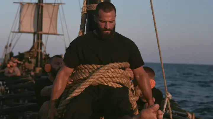 medium shot of a broad-shouldered Caucasian man in his early thirties with a square jaw, a short dark beard, a dark buzz cut and heavy brows, the build of a man who lifts, in a plain black t-shirt and black trousers,…