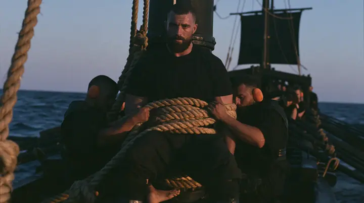 medium shot of a broad-shouldered Caucasian man in his early thirties with a square jaw, a short dark beard, a dark buzz cut and heavy brows, the build of a man who lifts, in a plain black t-shirt and black trousers,…