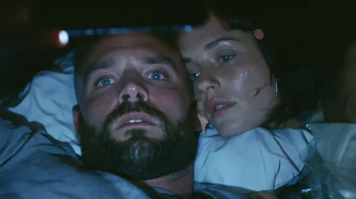 close-up of a broad-shouldered Caucasian man in his early thirties with a shaved head, a thick short dark beard, a square jaw, heavy brows and pale blue eyes, the build of a man who lifts, in a grey t-shirt, lying in…