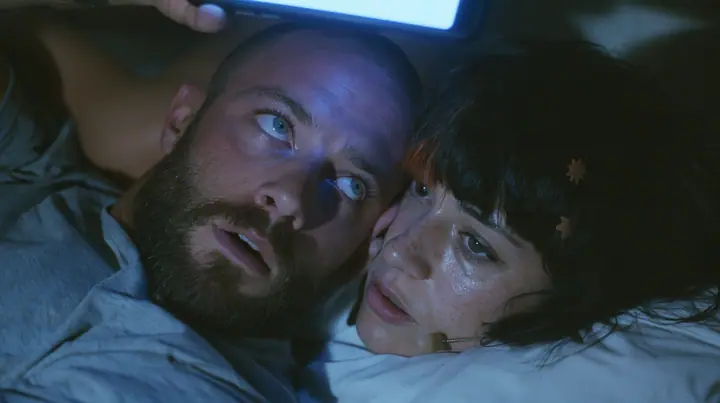 close-up of a broad-shouldered Caucasian man in his early thirties with a shaved head, a thick short dark beard, a square jaw, heavy brows and pale blue eyes, the build of a man who lifts, in a grey t-shirt, lying in…