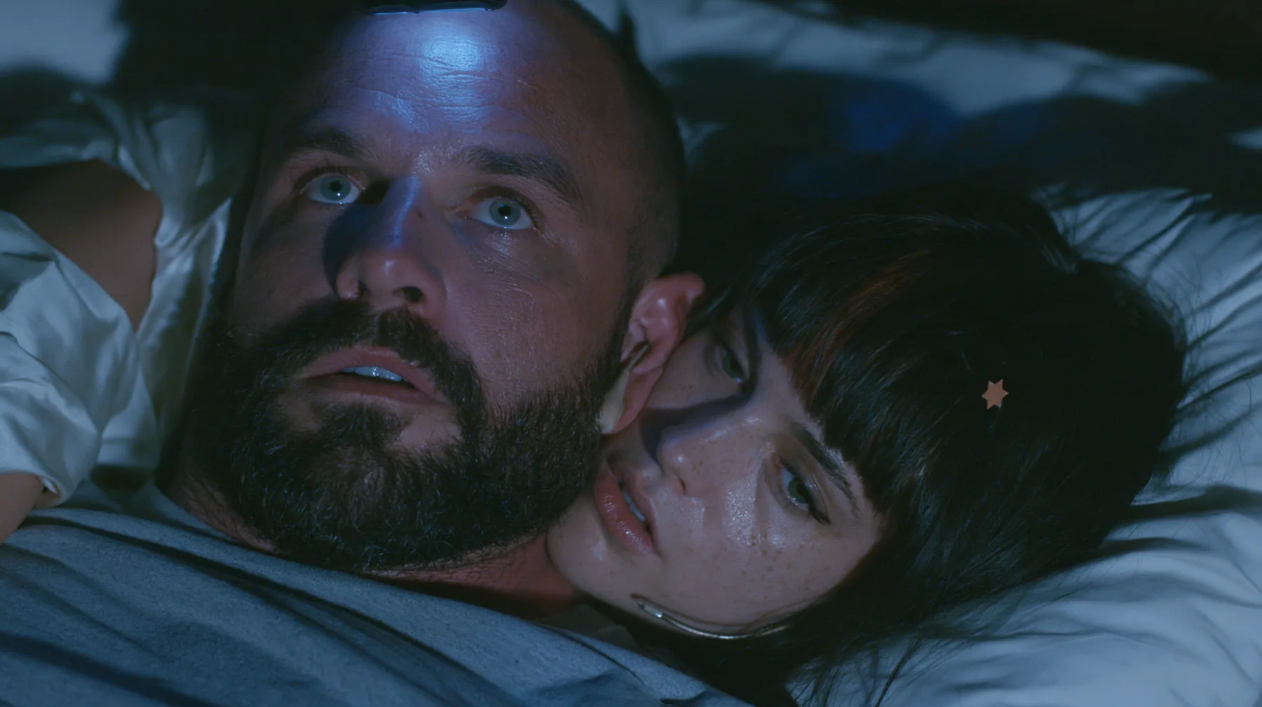 close-up of a broad-shouldered Caucasian man in his early thirties with a shaved head, a thick short dark beard, a square jaw, heavy brows and pale blue eyes, the build of a man who lifts, in a grey t-shirt, lying in…