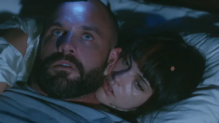 close-up of a broad-shouldered Caucasian man in his early thirties with a shaved head, a thick short dark beard, a square jaw, heavy brows and pale blue eyes, the build of a man who lifts, in a grey t-shirt, lying in…