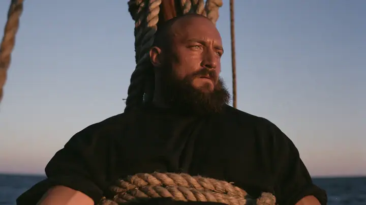 medium close-up of a broad-shouldered Caucasian man in his early thirties with a shaved head, a thick short dark beard, a square jaw, heavy brows and pale blue eyes, the build of a man who lifts, in a plain black…