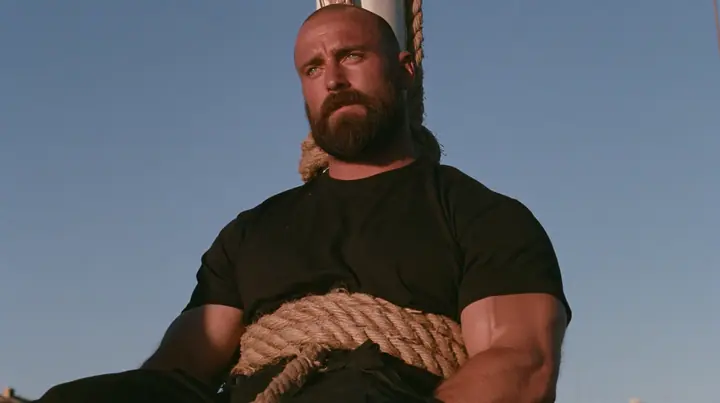 medium close-up of a broad-shouldered Caucasian man in his early thirties with a shaved head, a thick short dark beard, a square jaw, heavy brows and pale blue eyes, the build of a man who lifts, in a plain black…