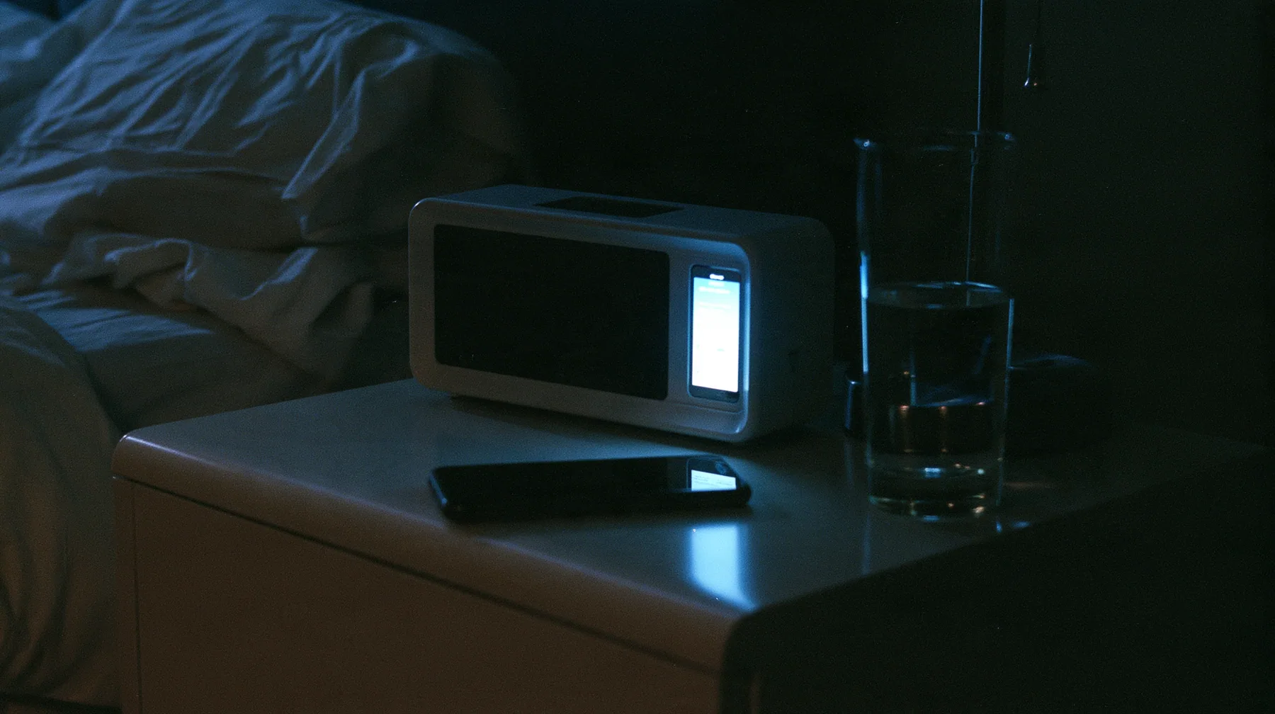 a bedside table in the dark with a blank black clock display, a glass of water and a phone face down, a dark bedroom at 3am lit only by a phone screen, blue-white glow on the face, cinematic 35mm film still, grain,…