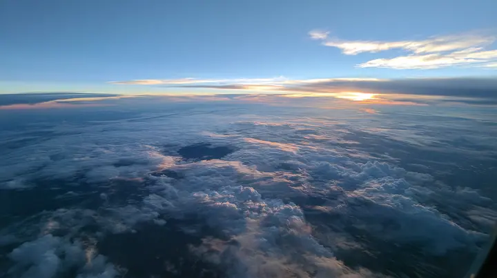 extreme wide aerial photograph from twelve kilometres above a white cloud deck at blue hour, one dark round gap in the clouds far below, the curve of the earth, no text, no letters, no logos
