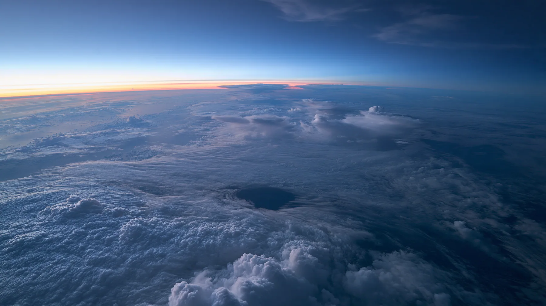 extreme wide aerial photograph from twelve kilometres above a white cloud deck at blue hour, one dark round gap in the clouds far below, the curve of the earth, no text, no letters, no logos