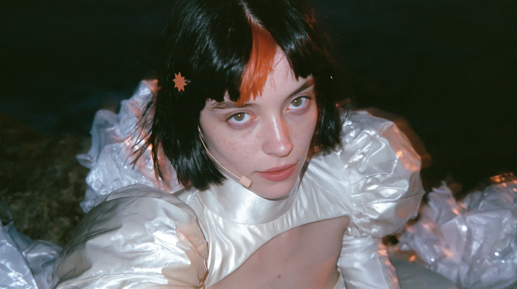 selfie-angle close-up of a Caucasian American woman in her mid-twenties with pale skin and light freckles, a glossy black blunt jaw-length bob with heavy straight bangs and one clay-orange streak through the bangs, a…