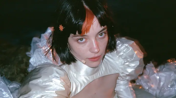 selfie-angle close-up of a Caucasian American woman in her mid-twenties with pale skin and light freckles, a glossy black blunt jaw-length bob with heavy straight bangs and one clay-orange streak through the bangs, a…
