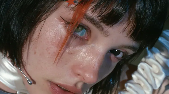extreme close-up of the right eye of a Caucasian American woman in her mid-twenties with pale skin and light freckles, a glossy black blunt jaw-length bob with heavy straight bangs and one clay-orange streak through…