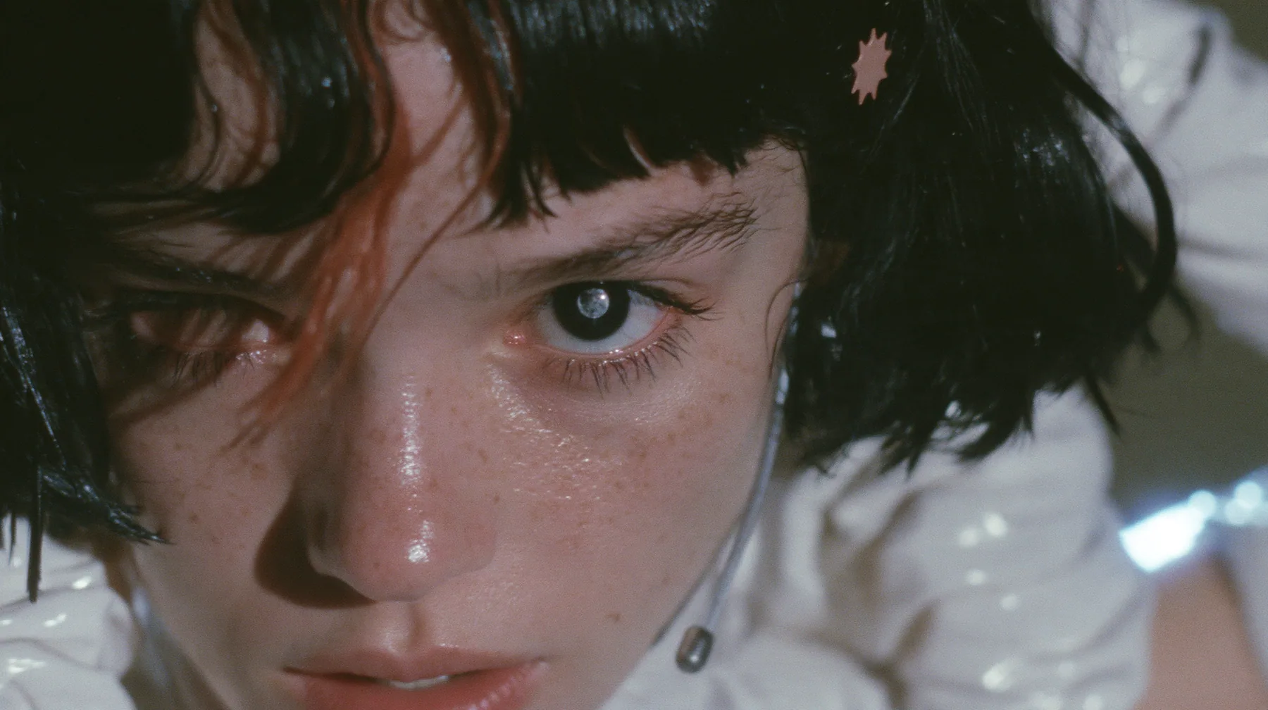 extreme close-up of the right eye of a Caucasian American woman in her mid-twenties with pale skin and light freckles, a glossy black blunt jaw-length bob with heavy straight bangs and one clay-orange streak through…
