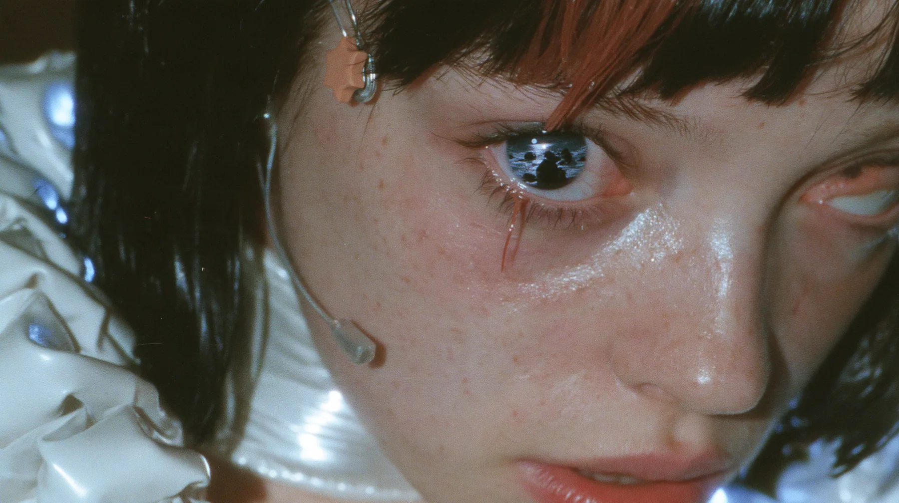 extreme close-up of the right eye of a Caucasian American woman in her mid-twenties with pale skin and light freckles, a glossy black blunt jaw-length bob with heavy straight bangs and one clay-orange streak through…