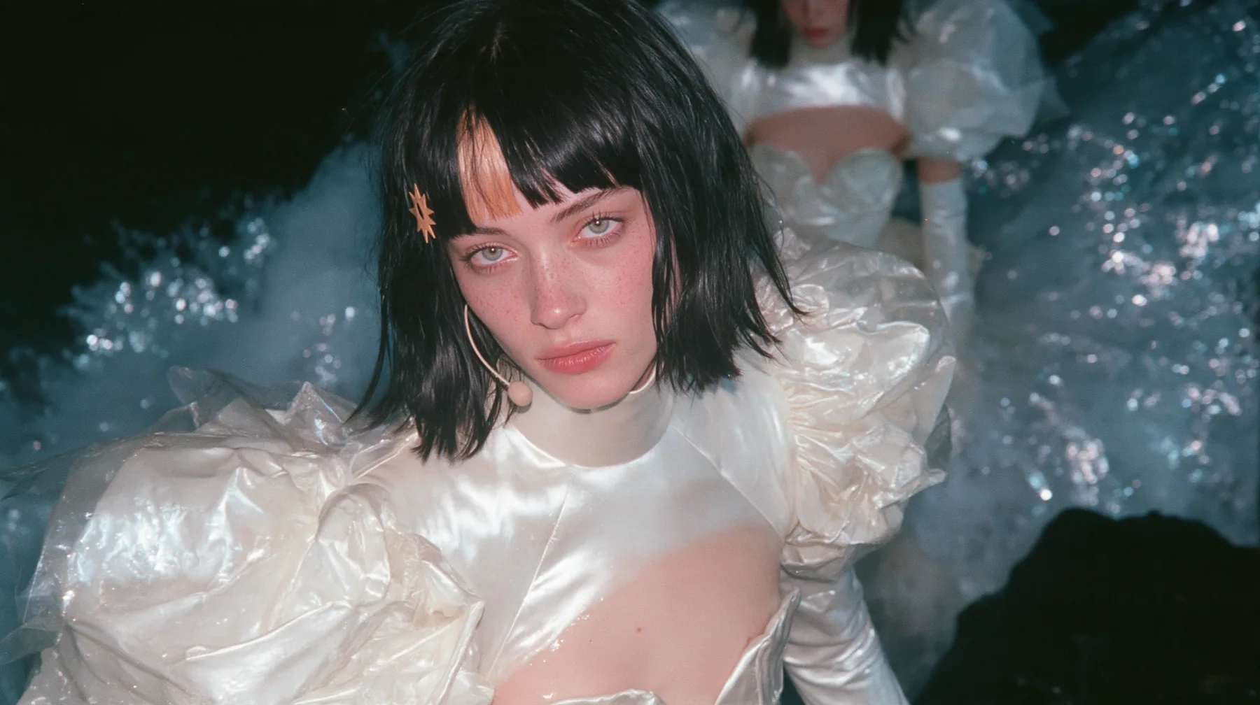 wide shot of a Caucasian American woman in her mid-twenties with pale skin and light freckles, a glossy black blunt jaw-length bob with heavy straight bangs and one clay-orange streak through the bangs, a small flat…