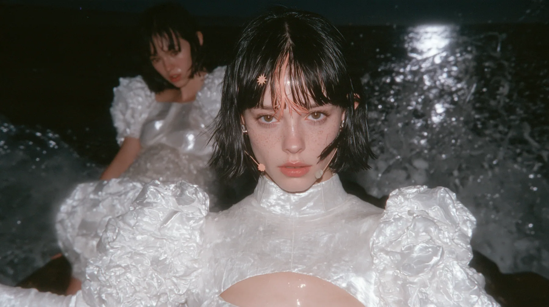 wide shot of a Caucasian American woman in her mid-twenties with pale skin and light freckles, a glossy black blunt jaw-length bob with heavy straight bangs and one clay-orange streak through the bangs, a small flat…