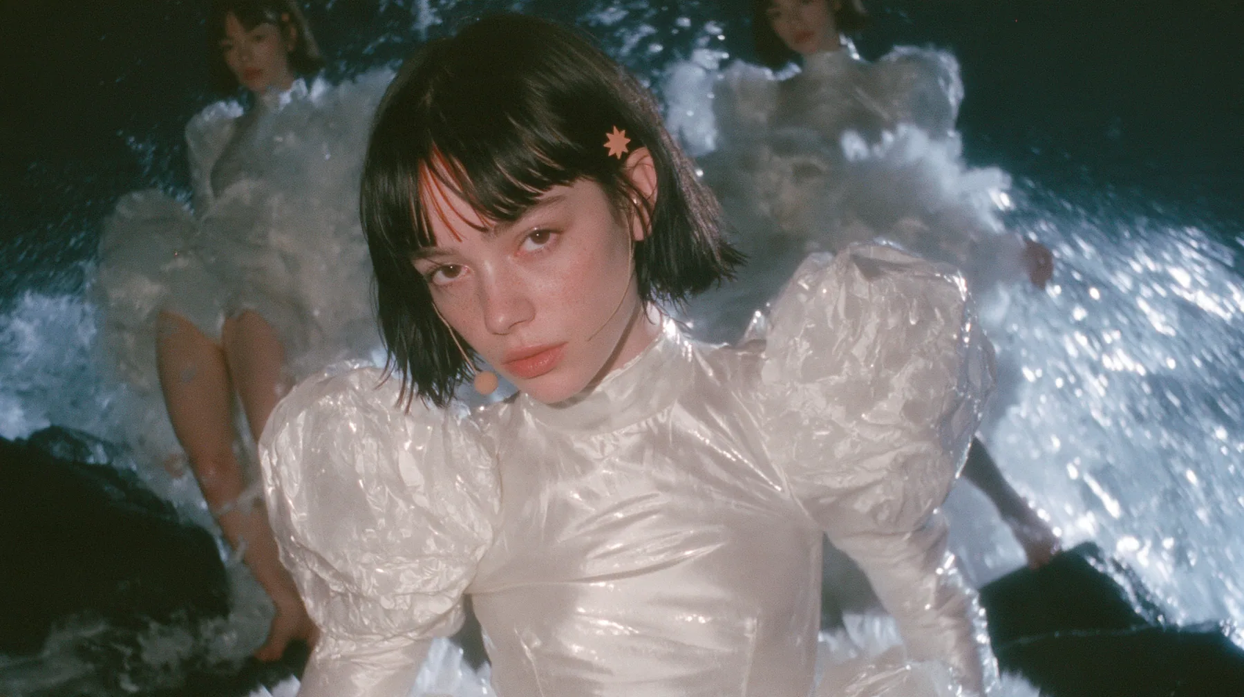 wide shot of a Caucasian American woman in her mid-twenties with pale skin and light freckles, a glossy black blunt jaw-length bob with heavy straight bangs and one clay-orange streak through the bangs, a small flat…