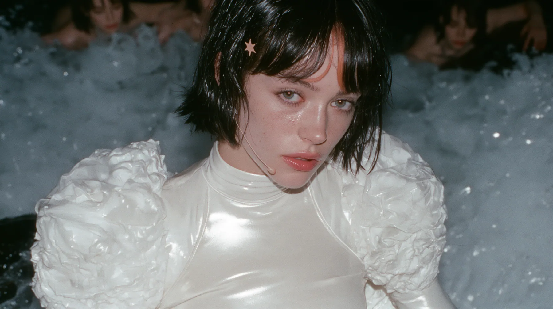 wide shot of a Caucasian American woman in her mid-twenties with pale skin and light freckles, a glossy black blunt jaw-length bob with heavy straight bangs and one clay-orange streak through the bangs, a small flat…
