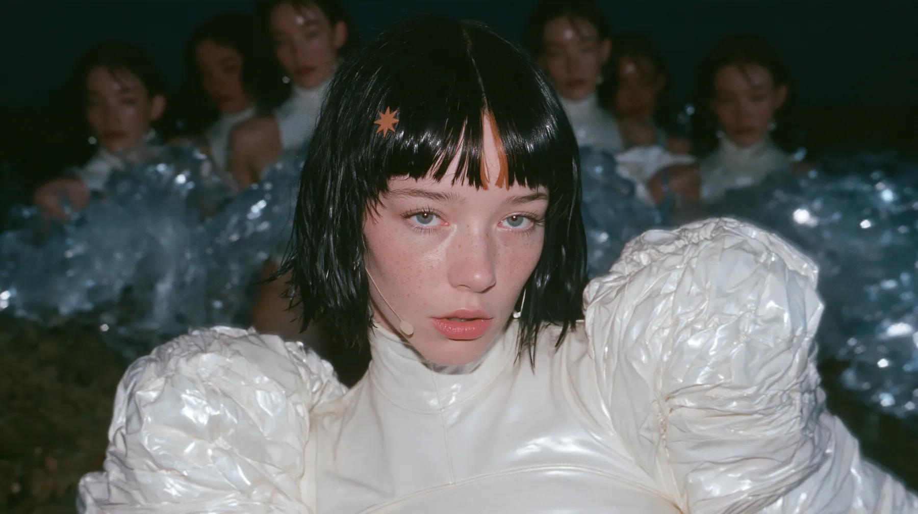 wide shot of a Caucasian American woman in her mid-twenties with pale skin and light freckles, a glossy black blunt jaw-length bob with heavy straight bangs and one clay-orange streak through the bangs, a small flat…