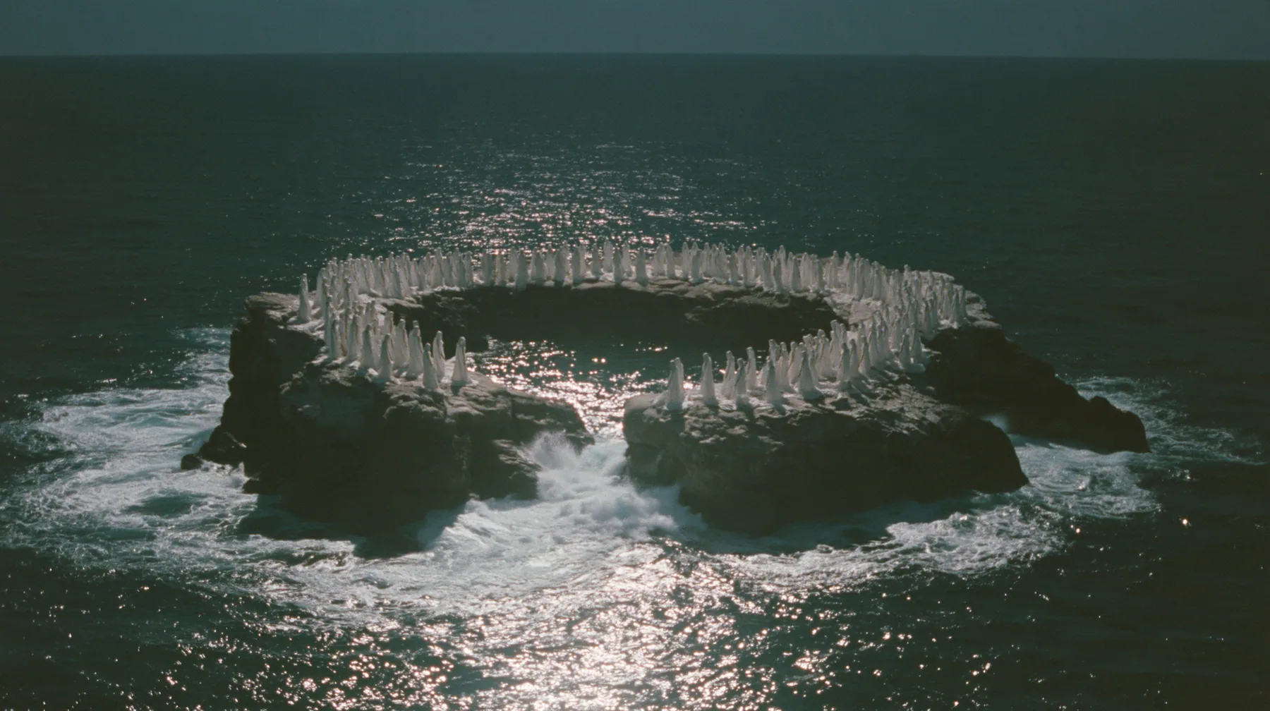 high wide aerial of a ring of black sea stacks covered with hundreds of identical pale women in white gowns, all facing the same direction, night, moonlight on a black-teal sea, silver spray, cinematic 35mm film…