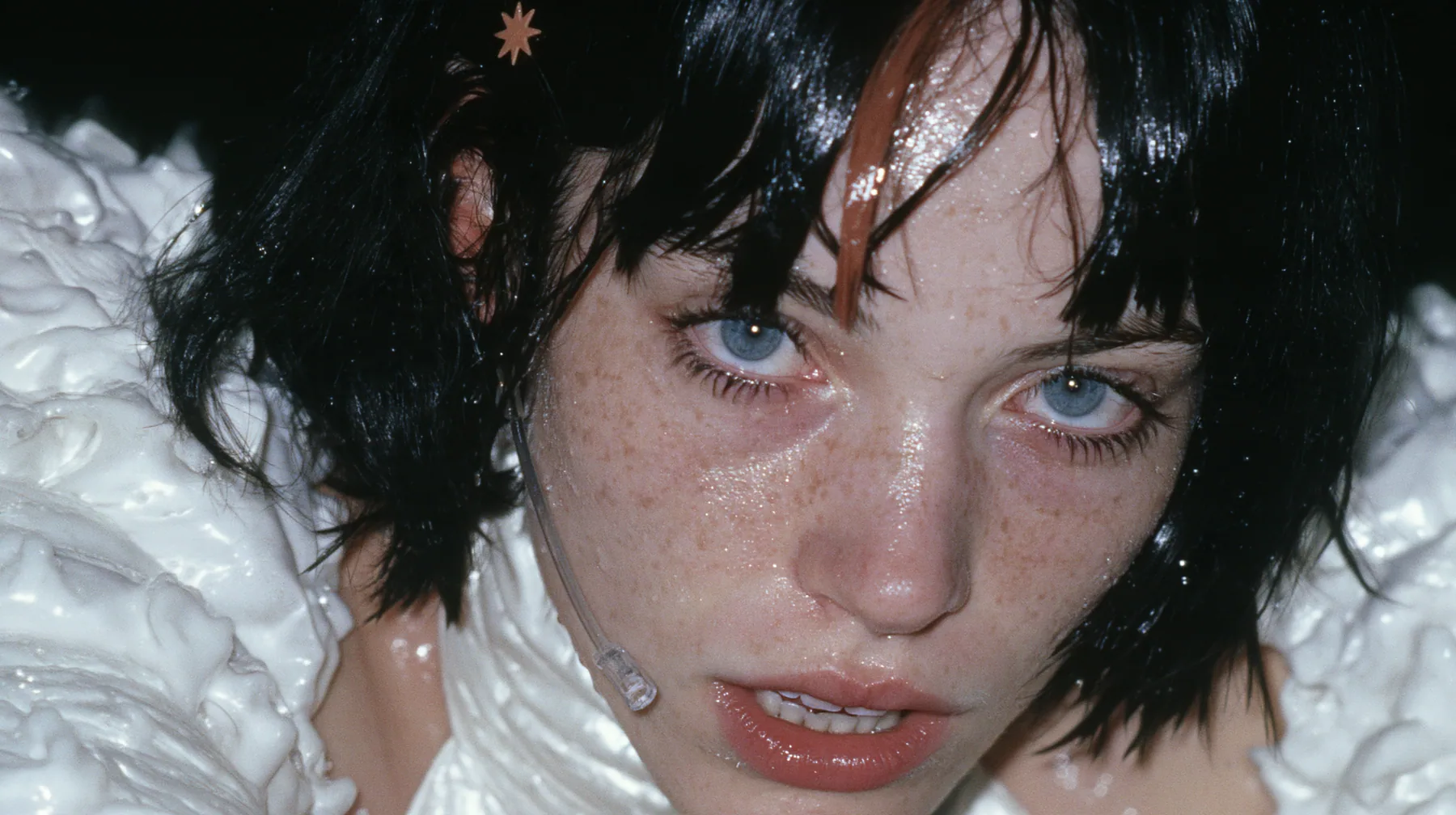 extreme close-up of the lips of a Caucasian American woman in her mid-twenties with pale skin and light freckles, a glossy black blunt jaw-length bob with heavy straight bangs and one clay-orange streak through the…