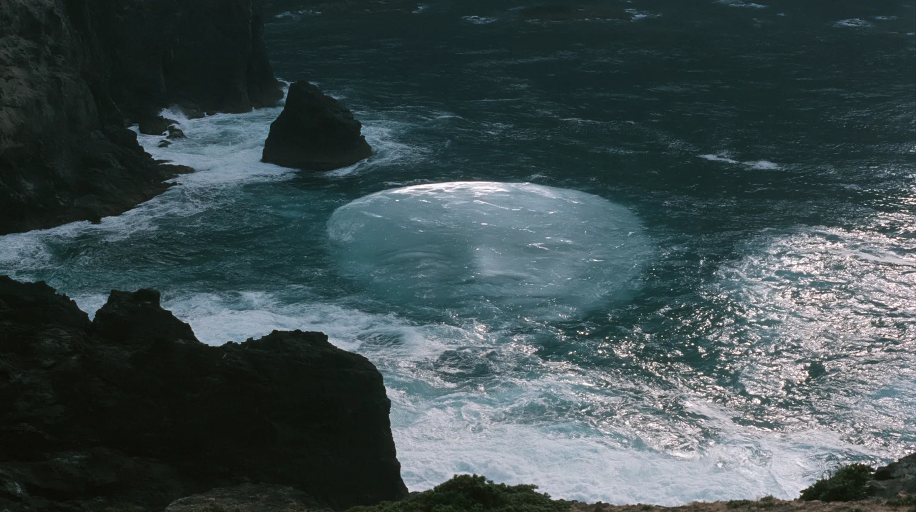 aerial photograph of a dark sea beside black rocks, just beneath the surface a colossal pale smooth oval shape like a face, barely visible, moonlight on the water, night, moonlight on a black-teal sea, silver spray,…