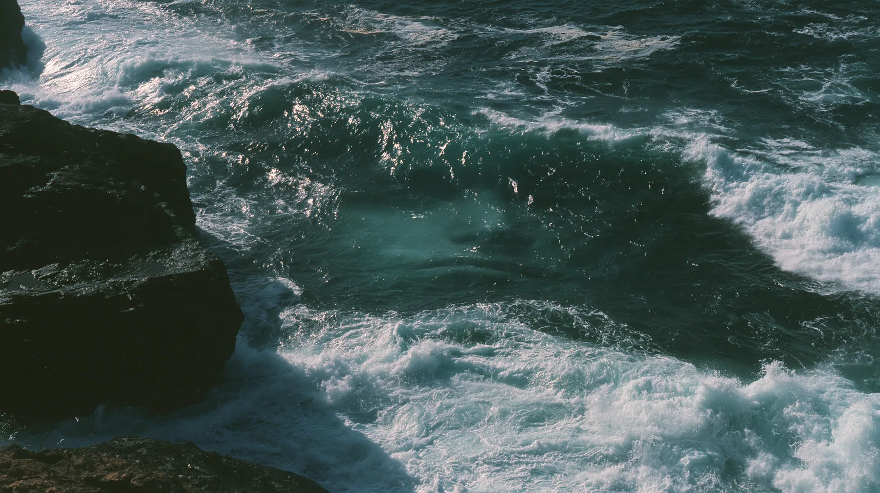 aerial photograph of a dark sea beside black rocks, just beneath the surface a colossal pale smooth oval shape like a face, barely visible, moonlight on the water, night, moonlight on a black-teal sea, silver spray,…