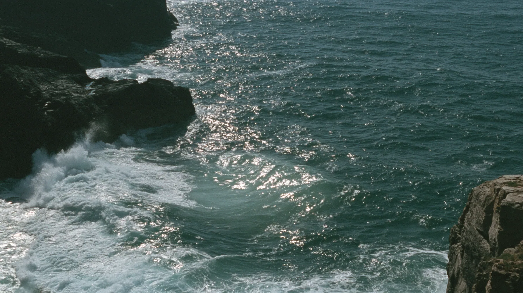 aerial photograph of a dark sea beside black rocks, just beneath the surface a colossal pale smooth oval shape like a face, barely visible, moonlight on the water, night, moonlight on a black-teal sea, silver spray,…