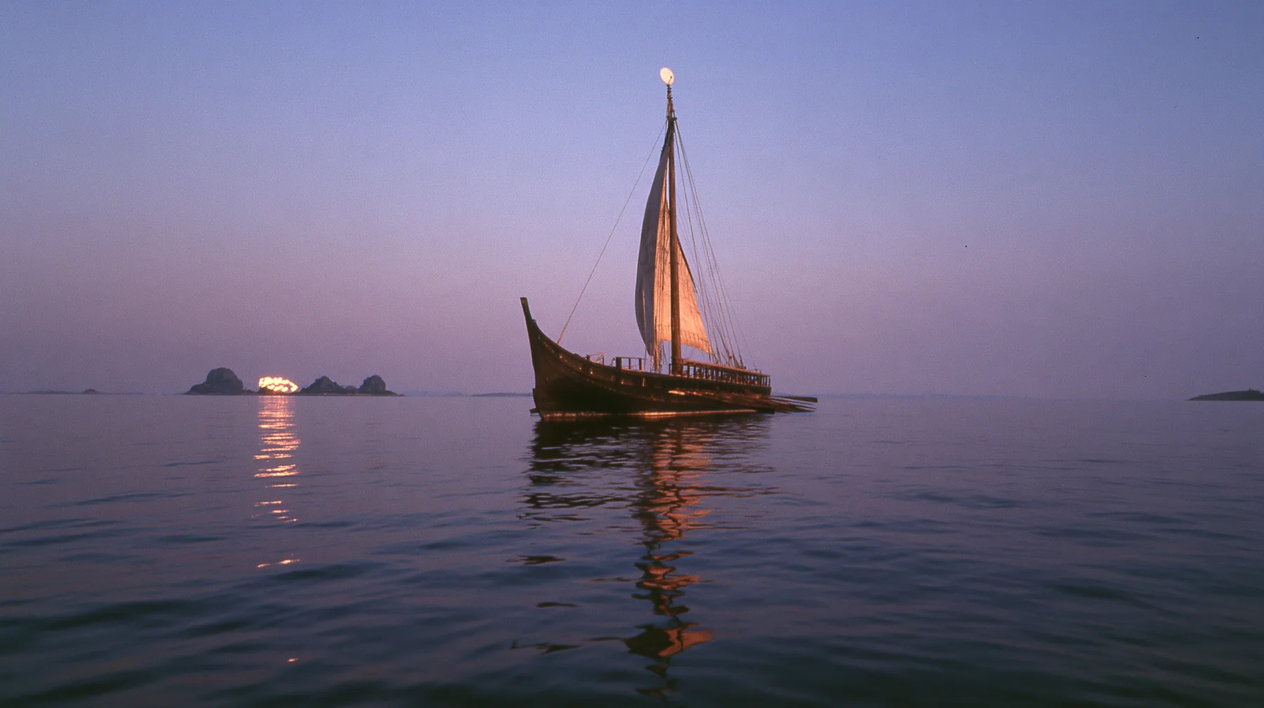 low angle up the mast of a long black ancient Greek galley with a single mast, a square sail and two banks of oars, a small white satellite dish at the top, distant rocks glowing gold on the horizon, dusk, low gold…