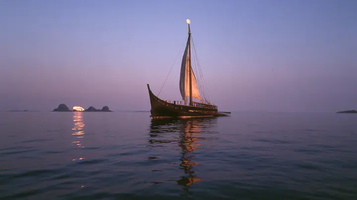 low angle up the mast of a long black ancient Greek galley with a single mast, a square sail and two banks of oars, a small white satellite dish at the top, distant rocks glowing gold on the horizon, dusk, low gold…