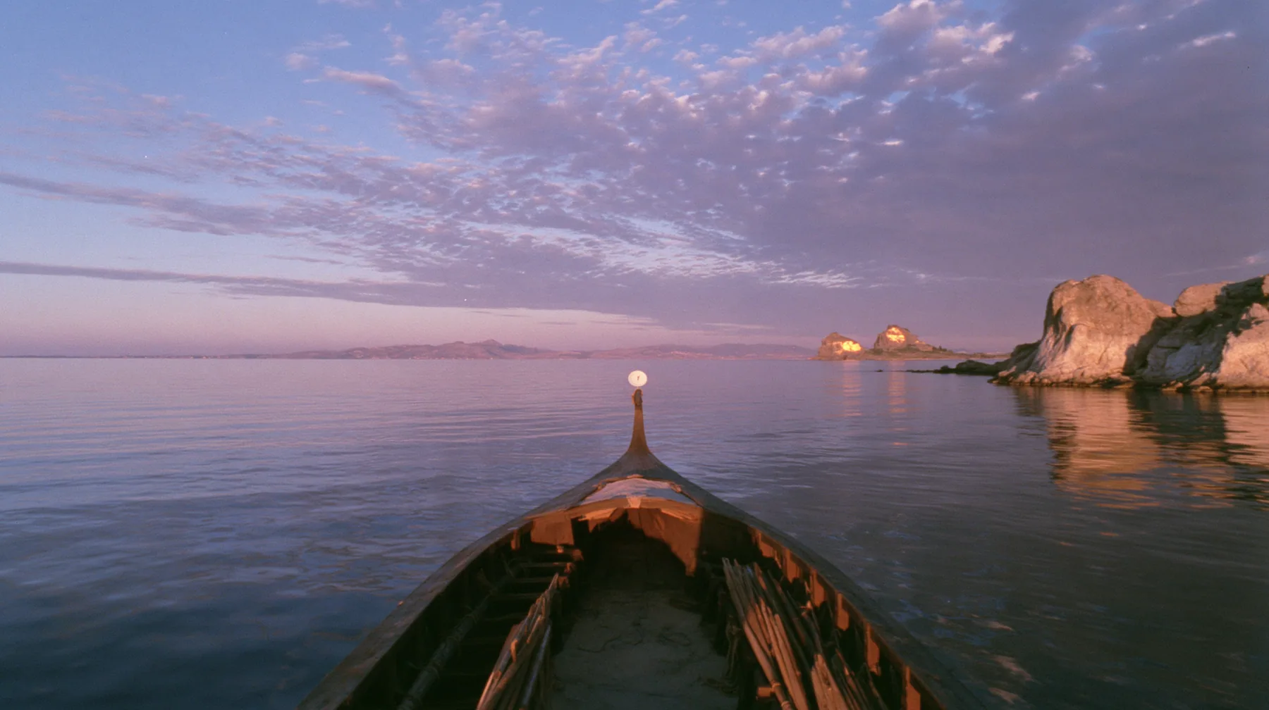 low angle up the mast of a long black ancient Greek galley with a single mast, a square sail and two banks of oars, a small white satellite dish at the top, distant rocks glowing gold on the horizon, dusk, low gold…