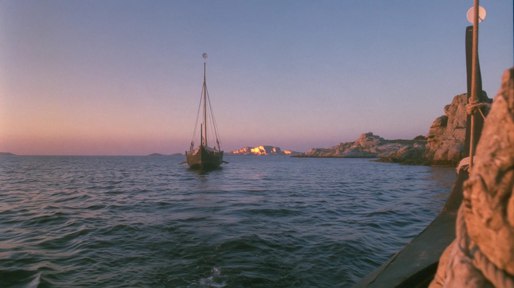 low angle up the mast of a long black ancient Greek galley with a single mast, a square sail and two banks of oars, a small white satellite dish at the top, distant rocks glowing gold on the horizon, dusk, low gold…